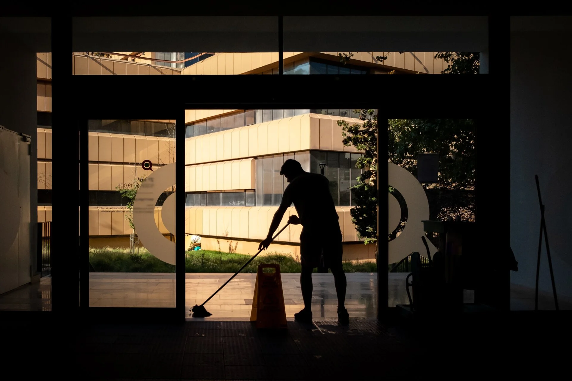 Silhouette of a person sweeping a wet courtyard floor with a broom and dustpan, backlit by warm sunlight streaming through glass doors and windows of a modern building with yellow-toned architecture.