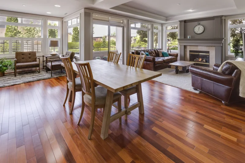 Spacious open-concept living room featuring rich medium-brown hardwood flooring, dining table with wooden chairs, brown leather seating, fireplace, and expansive windows overlooking outdoor landscape.