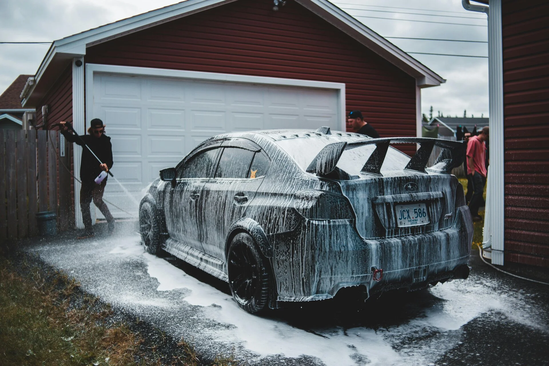 A dark-colored SUV covered in soapy water streams during an outdoor car wash in a residential driveway. Multiple people in casual clothing spray and clean the vehicle with high-pressure water in front of a red-brown garage with white trim.