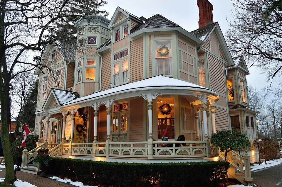 A pale green Victorian mansion with ornate white porch columns and railings, decorated with golden wreaths and garland. Warm interior lights glow through multiple windows, and snow covers the ground around the home surrounded by bare trees.