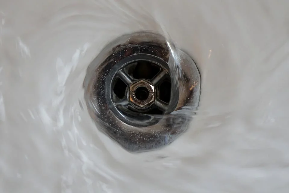 Close-up view of a white porcelain sink drain with dark mineral deposits and buildup around the metal drain fixture and central opening, showing signs of age and mineral accumulation.