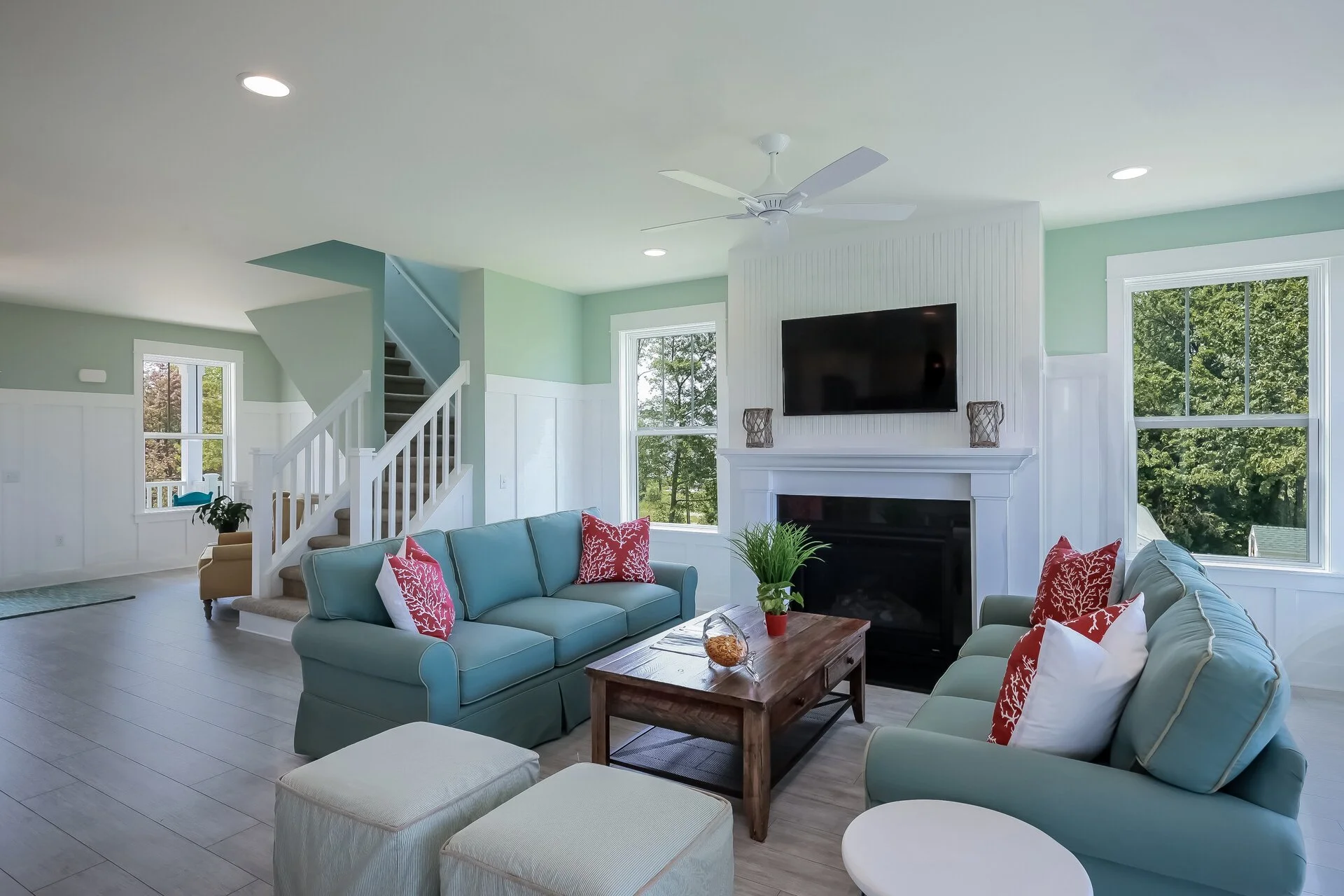 Bright, open-concept living room featuring mint green and white walls, turquoise upholstered seating with red patterned pillows, wooden coffee table, fireplace with flat-screen TV, white staircase, recessed lighting, ceiling fan, and large windows overlooking green trees.