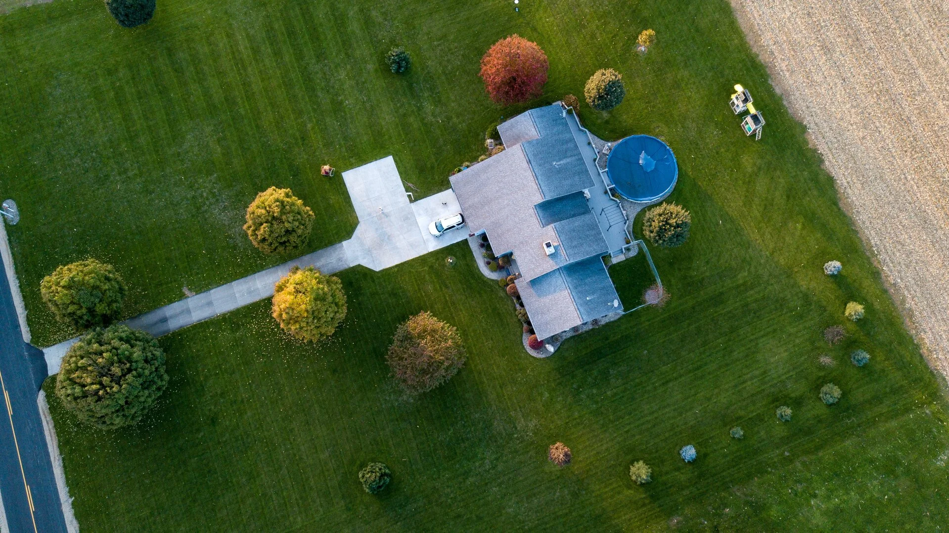Overhead view of a modern house with blue swimming pool surrounded by lush green lawn, scattered ornamental trees in various fall colors, driveway, and adjacent gravel area.