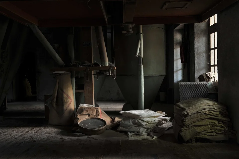 Dimly lit abandoned industrial space with scattered debris including metal containers, a cast iron pan, stacked fabric bundles, and deteriorating materials on concrete floor, with sunlight streaming through barred windows