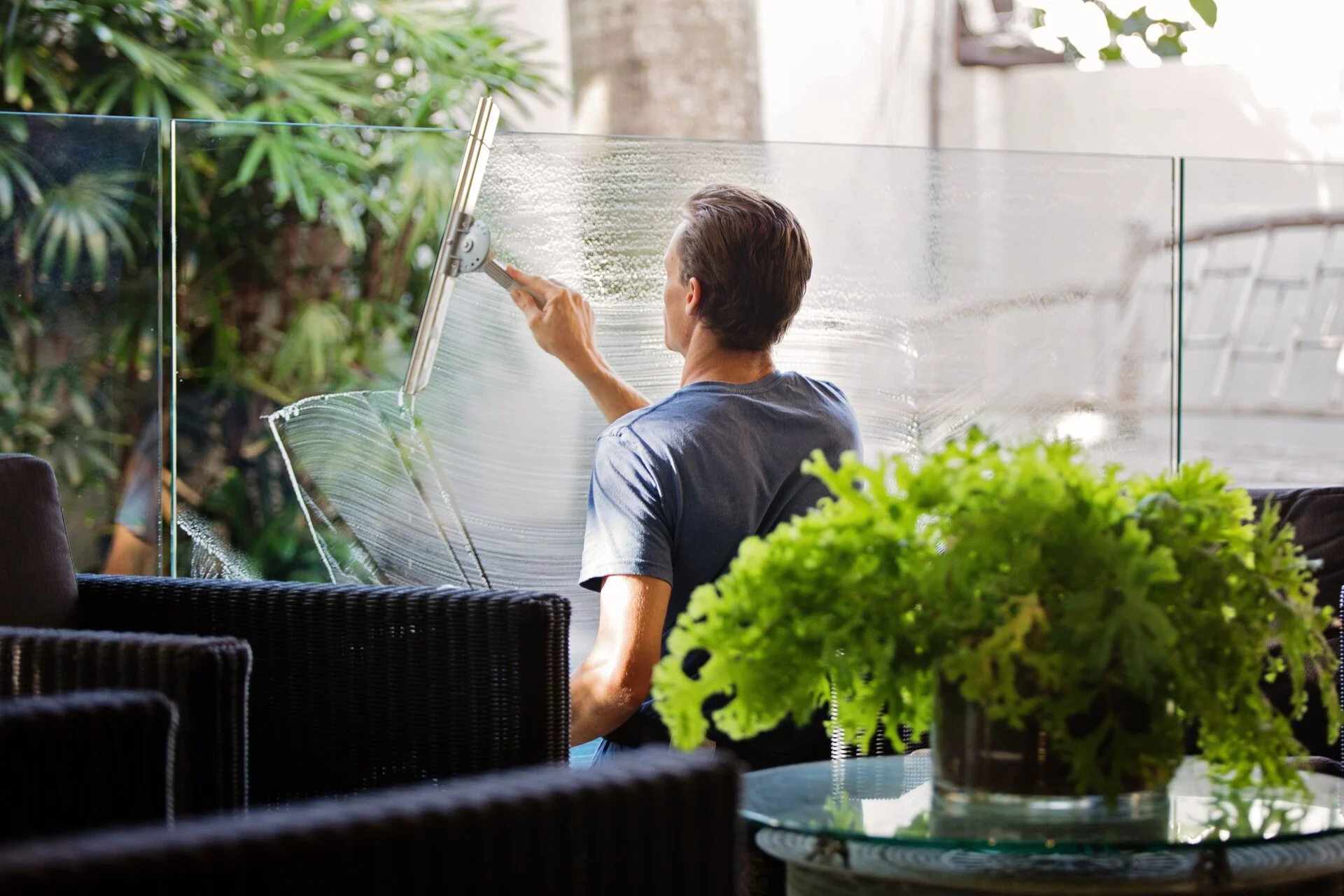 Man in blue shirt cleaning a large glass window with a squeegee tool on a sunny patio. Green potted plants and black wicker furniture visible in foreground, with building architecture in background.