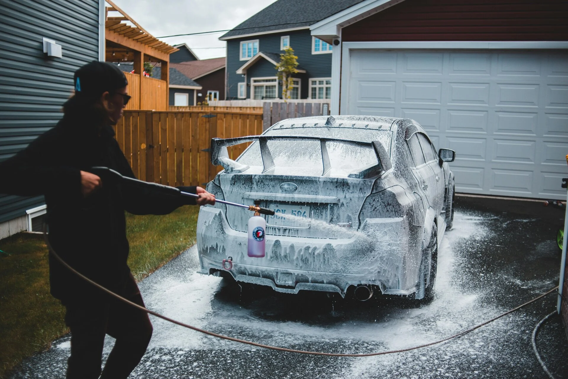 A person in black clothing uses a pressure washer to spray water on a white sedan in a residential driveway, with suburban houses and a garage visible in the background.