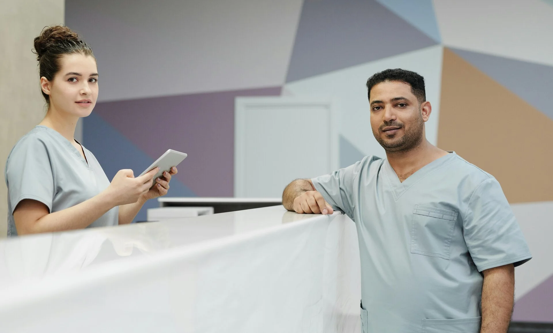 Female dental receptionist in light blue scrubs holding a tablet at a white reception desk, interacting with a male patient in matching scrubs in a modern dental office with geometric wall design.