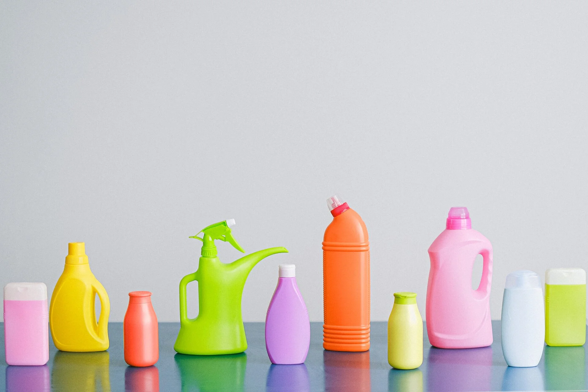 Row of ten cleaning supply bottles and containers in bright colors including pink, yellow, red, lime green, purple, orange, and blue arranged on a reflective gray surface against a light gray background.