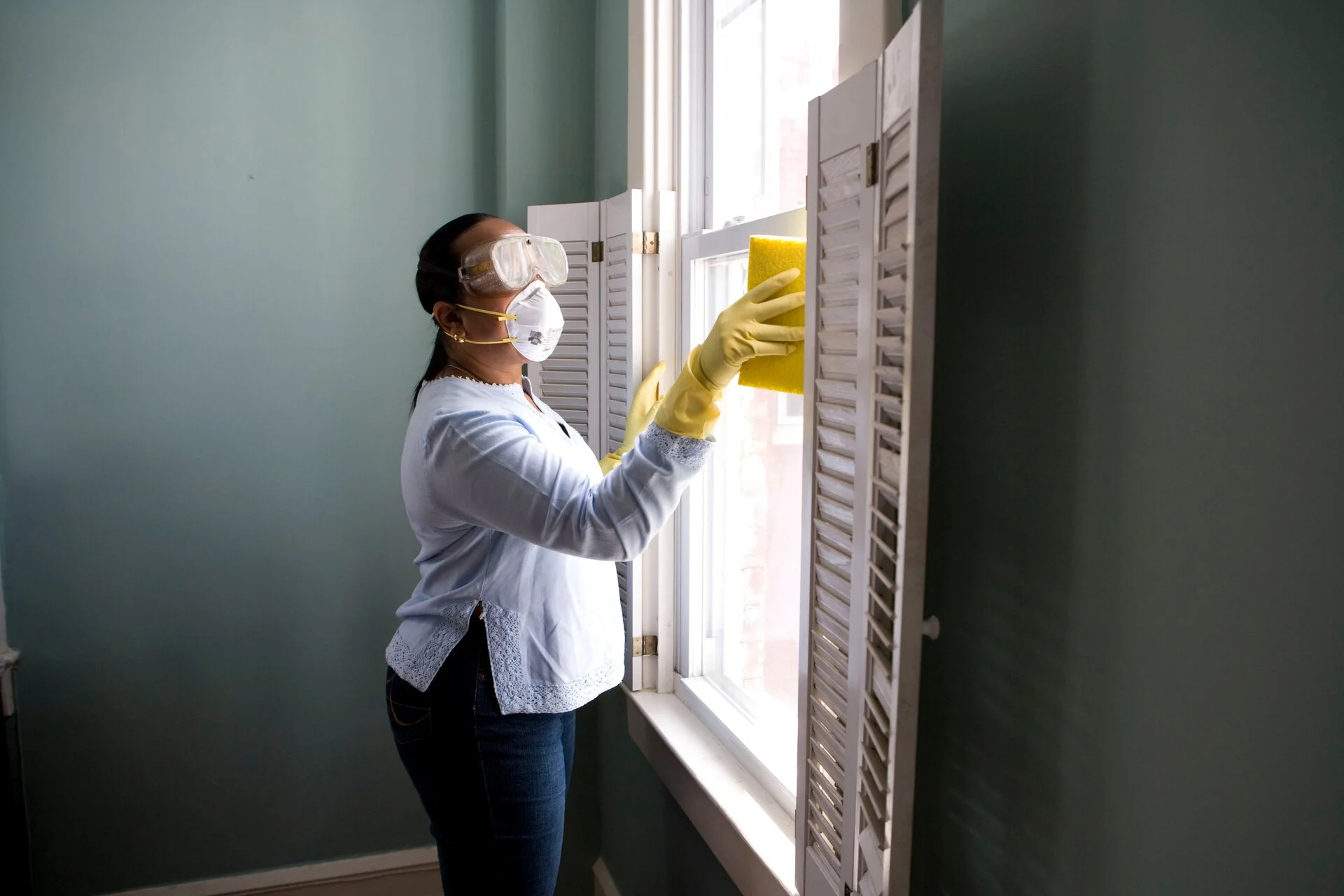 Woman wearing white protective mask, goggles, and yellow rubber gloves cleaning a bright window with white frame and shutters in a light blue room.