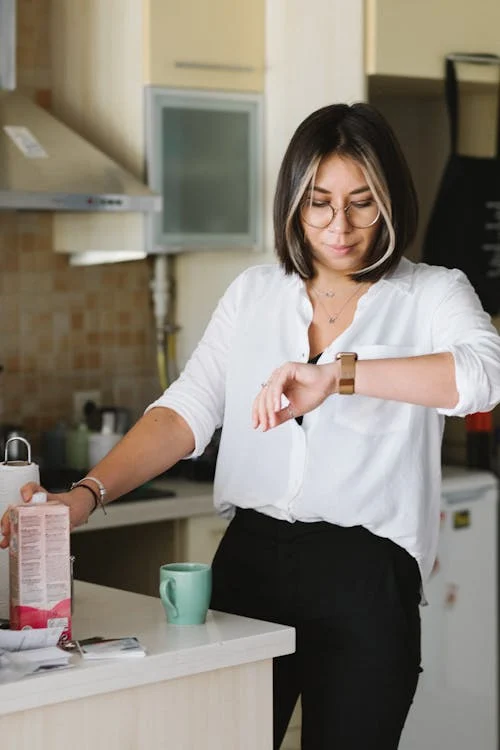 Woman wearing glasses and white shirt checks her gold wristwatch in a modern kitchen, with a green mug and items on the counter beside her.