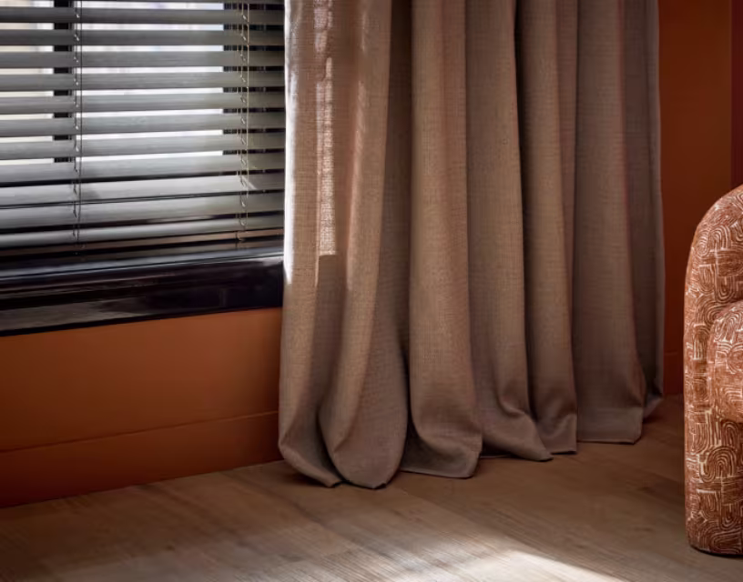Taupe gordijnen hangen naast een raam met zwarte, houten jaloezieën, zonlicht valt op een houten vloer met een decoratief kussen.