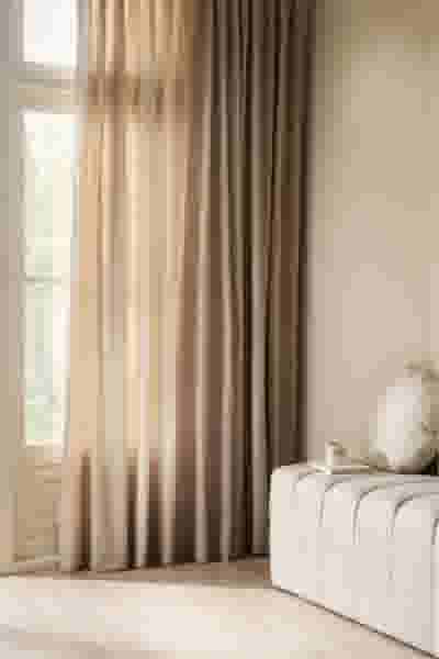 Lichtbeige gordijnen naast een raam in een rustige woonkamer met een witte bank en een decoratief kussen.