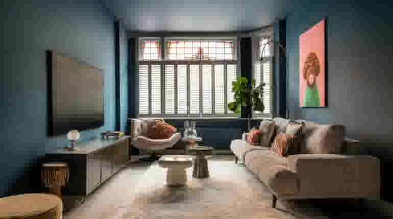 Moderne woonkamer met blauwe muren, beige bank, glas-in-loodramen, tv-meubel en kleurrijk kunstwerk aan de muur.