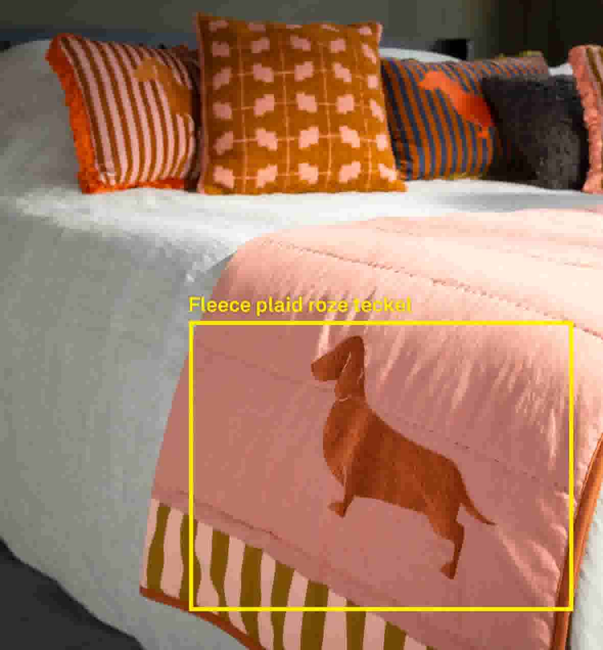 Sfeerbeeld van een bed met witte sprei, roze plaid met teckelmotief en olijfgroen-roze gestreepte rand, met sierkussens in gestreepte, geruite en effen stoffen op de achtergrond.
