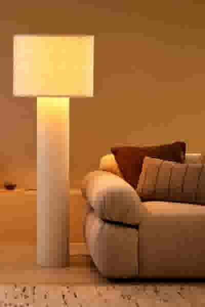 Staande lamp met crèmekleurige kap naast een beige bank met bruine kussens in een warm verlichte woonkamer.