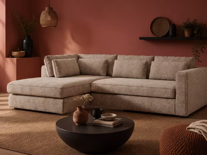 Beige hoekbank met chaise longue in warme woonkamer met terracotta muur, ronde zwarte salontafel en rotan vloerkleed.