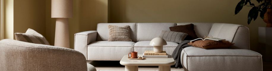 Moderne woonkamer met beige muren, grijze bank met donker kussen, witte vaas, vloerlamp en rode bijzettafel.
