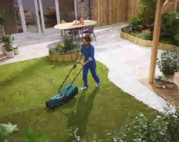 Vrouw in blauwe overall maait gras met groene grasmaaier in verzorgde tuin met terras en houten pergola.
