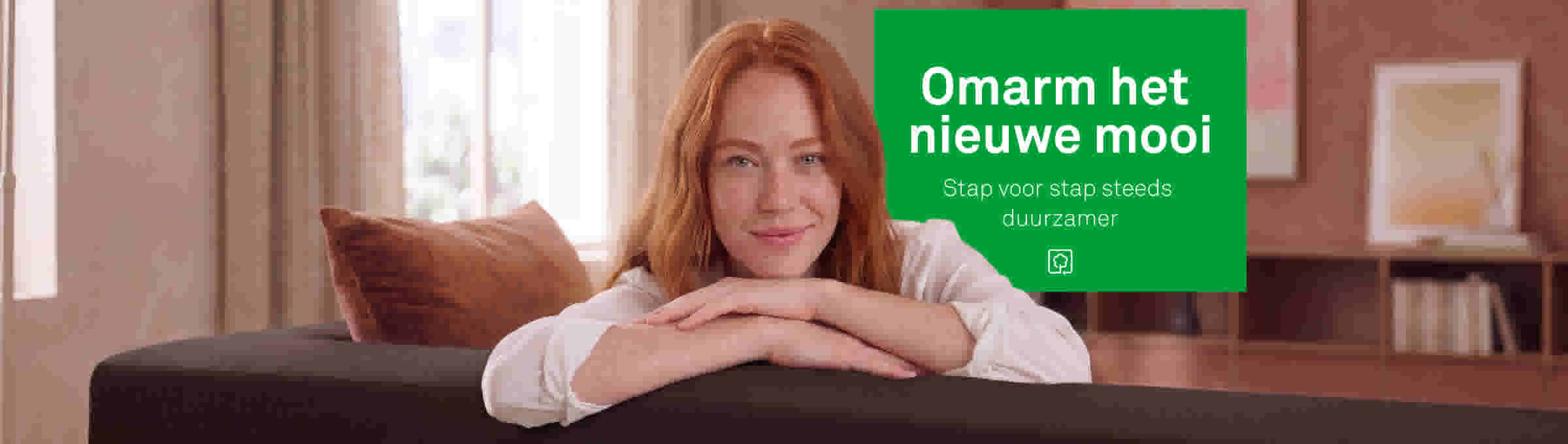 Vrouw met rood haar leunt glimlachend op een donkere bank in een warme, roze woonkamer met kunst aan de muur.