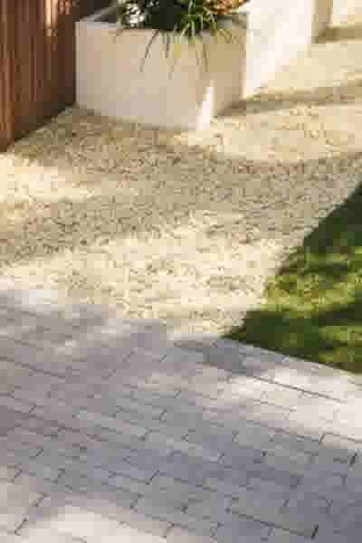 Grijze bestrating met rechthoekige stenen, omgeven door wit grind, groen gras en een witte plantenbak met groen.