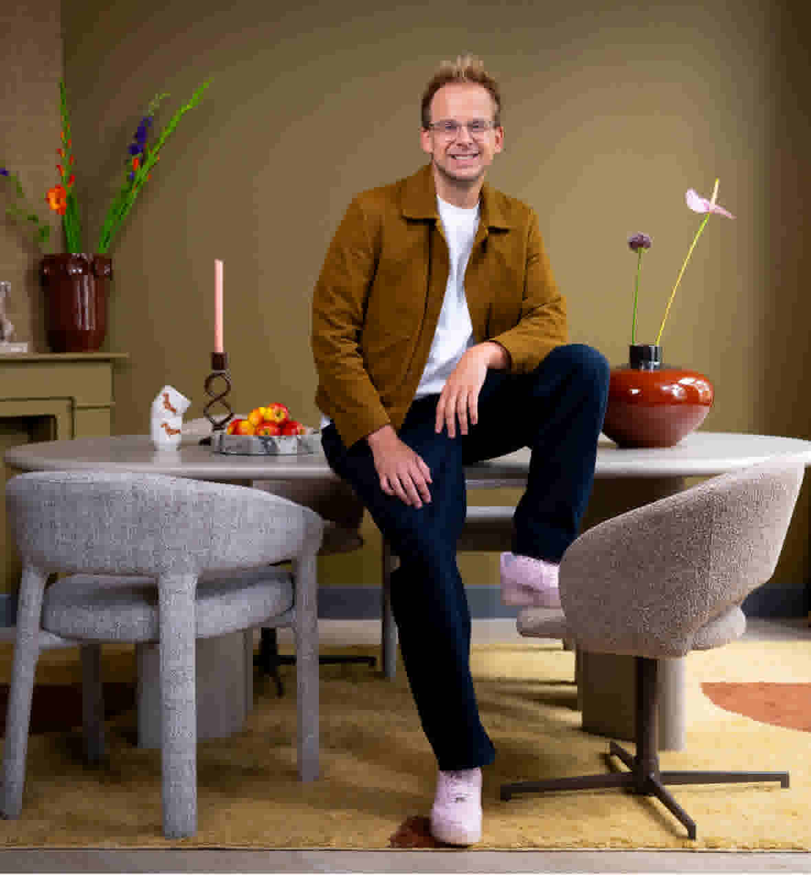 Sfeerbeeld van Theo-Bert Pot, zittend op een eettafel met grijze stoelen, met vazen bloemen, een schaal fruit en een kandelaar, in een kamer met olijfgroene wanden en een geel vloerkleed.