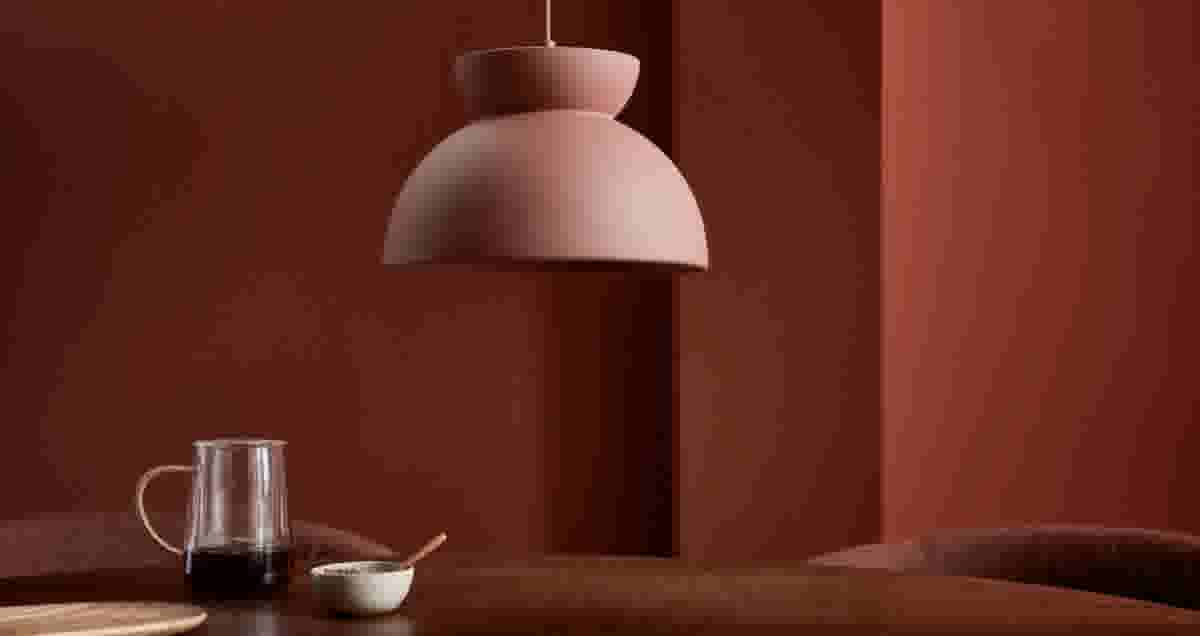Roze hanglamp boven een houten tafel met een glazen kan en een kommetje, tegen een terracotta rode muur.