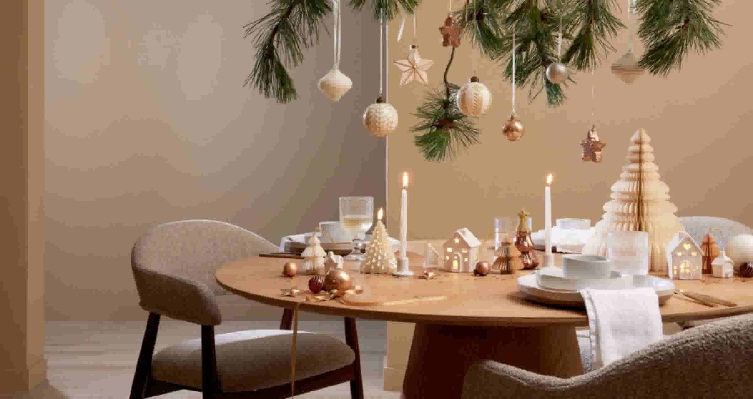 Feestelijk gedekte eettafel met kaarsen en kerstversiering, dennengroen met hangende ornamenten erboven in warme neutrale tinten.