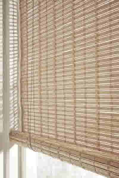 Lichtbruine bamboe rolgordijn aan een raam, gedeeltelijk opgerold, met zacht licht dat erdoorheen filtert.