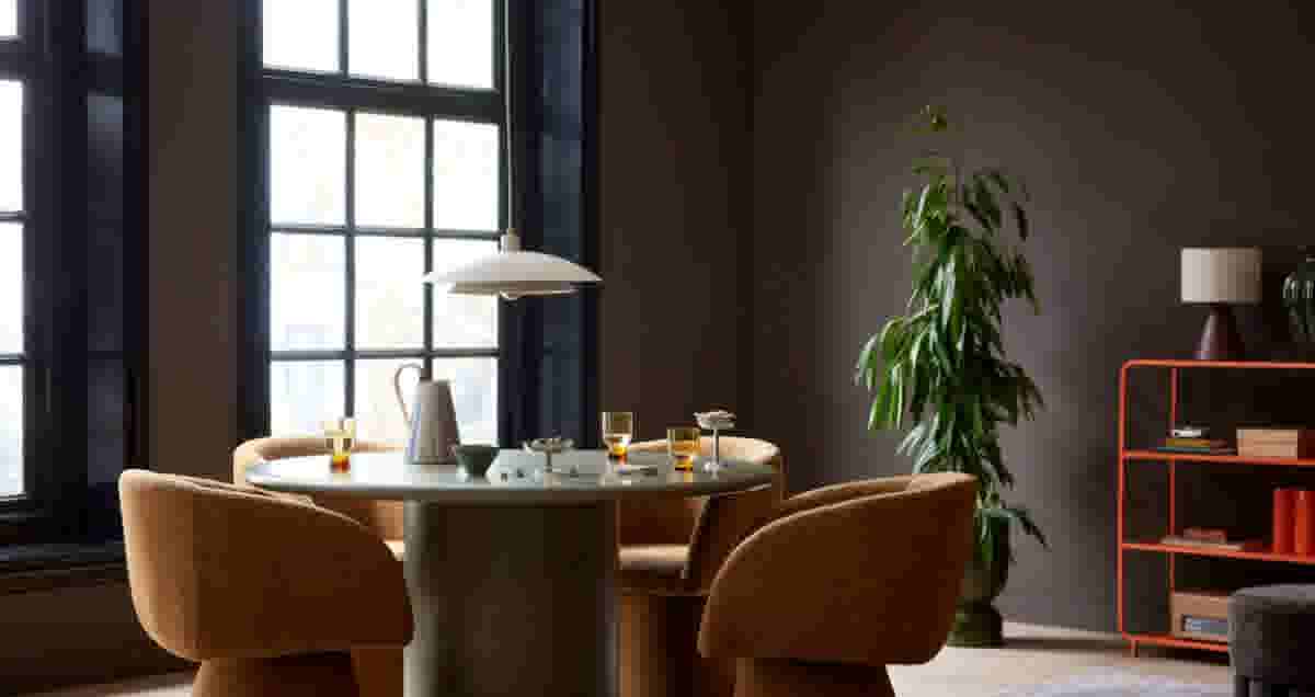Stijlvolle eetkamer met groene ronde tafel, bruine stoelen, hanglamp en plant. Oranje boekenkast en grote ramen op achtergrond.
