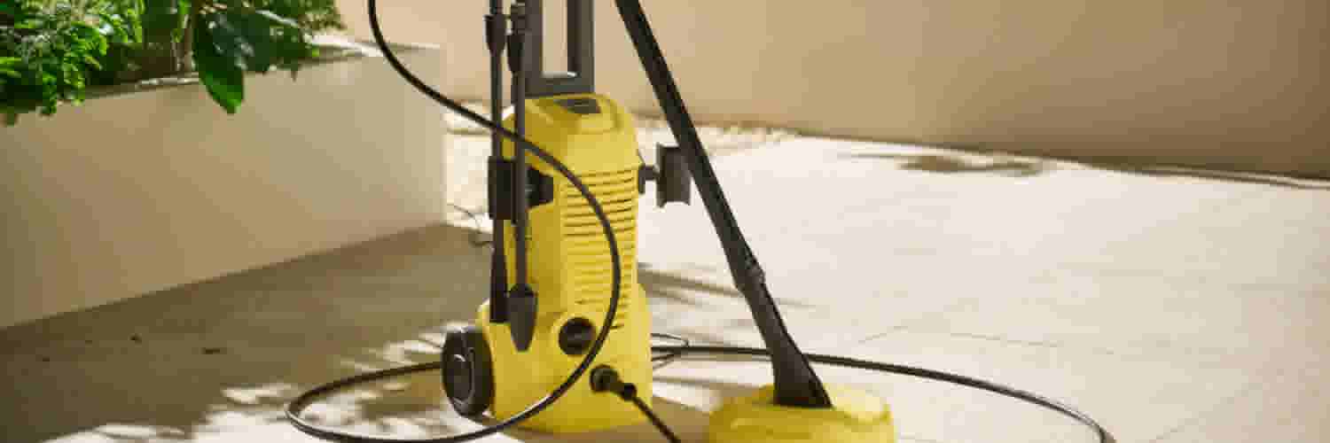 Gele hogedrukreiniger met zwarte slang op een betegelde vloer naast een groene plant in een witte pot.