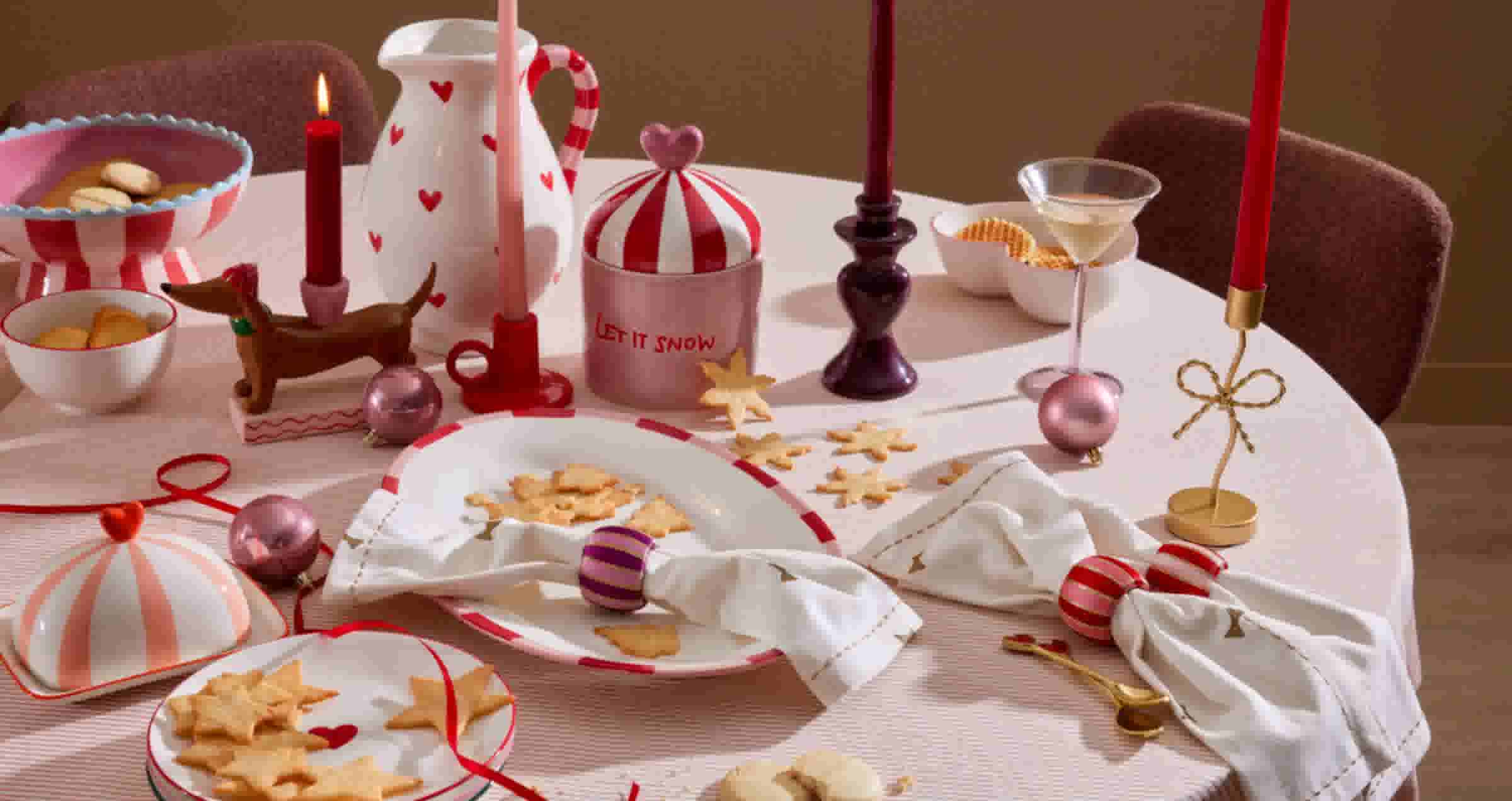 Feestelijk gedekte tafel met kerstversiering, rode kaarsen, een kan met hartjes en sterkoekjes op witte borden.