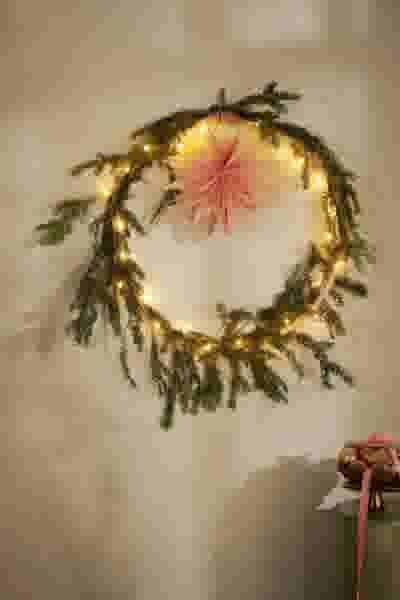 Feestelijke kerstkrans met dennentakken, lichtjes en een roze papieren ster, hangend aan een lichte muur naast een cadeau.