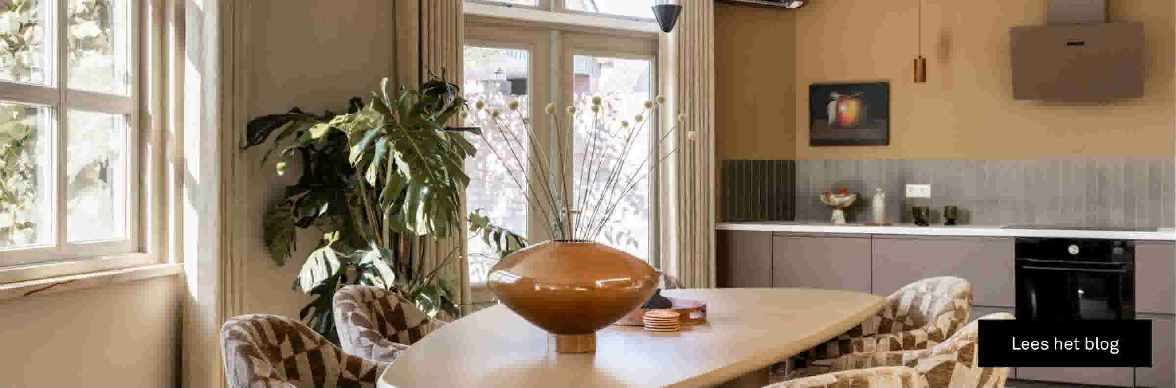 Lichte eetkamer met ronde tafel, gestoffeerde stoelen, grote ramen, kamerplanten en moderne keuken op de achtergrond.