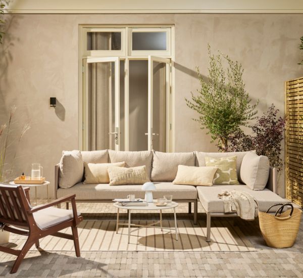 Gezellig terras met beige loungebank, houten stoel en groene planten tegen een lichte muur met openslaande deuren.