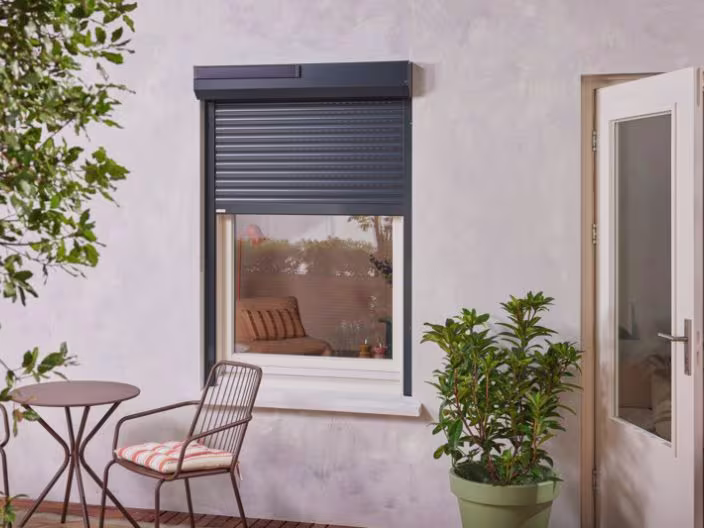 Moderne woning met donkerblauw rolluik, terras met stoel en tafel, groene planten en glazen deur aan de rechterkant.