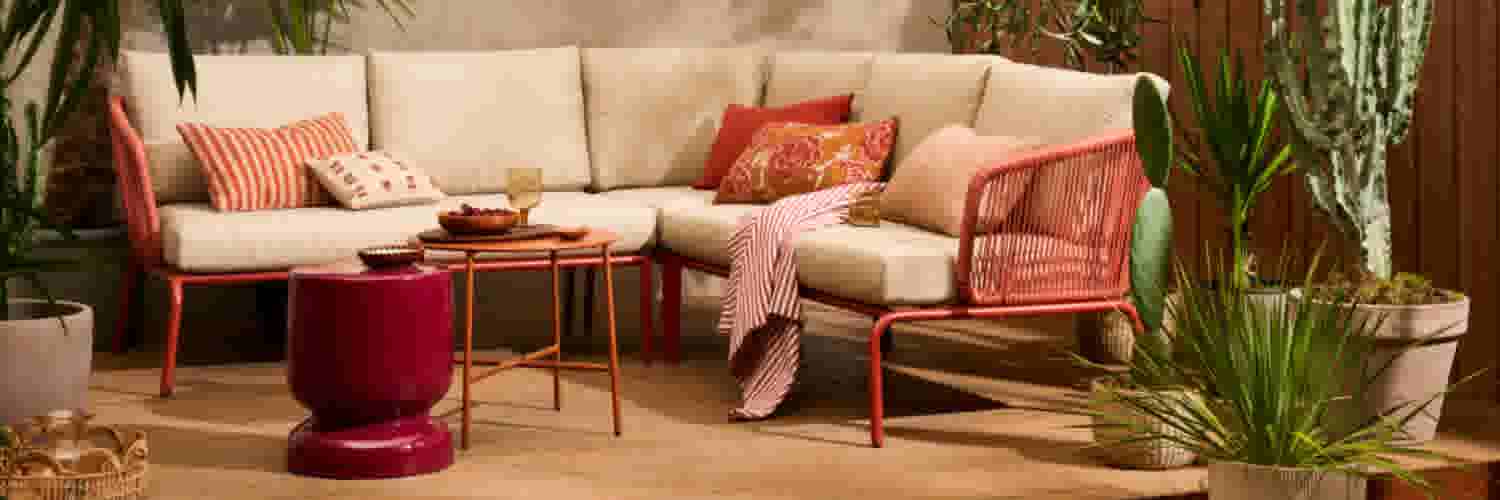 Gezellige buitenlounge met beige hoekbank, rode accenten en tropische planten op een houten terras.