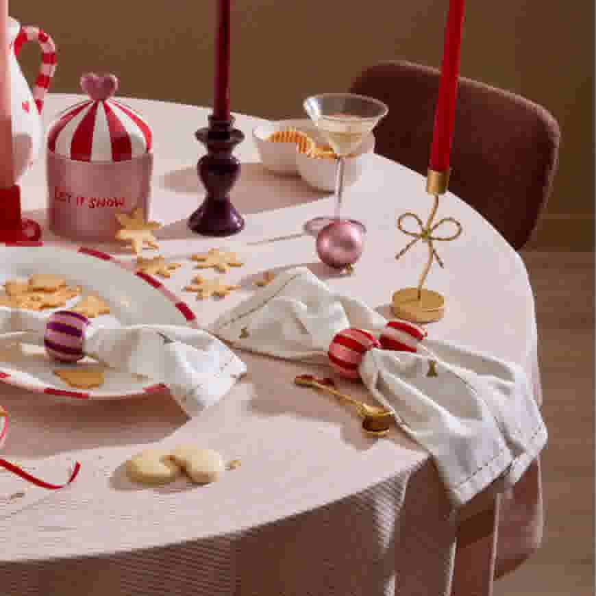 Feestelijk gedekte tafel met kerstversiering, koekjes, kaarsen en een cocktailglas op een wit-rood gestreept tafelkleed.