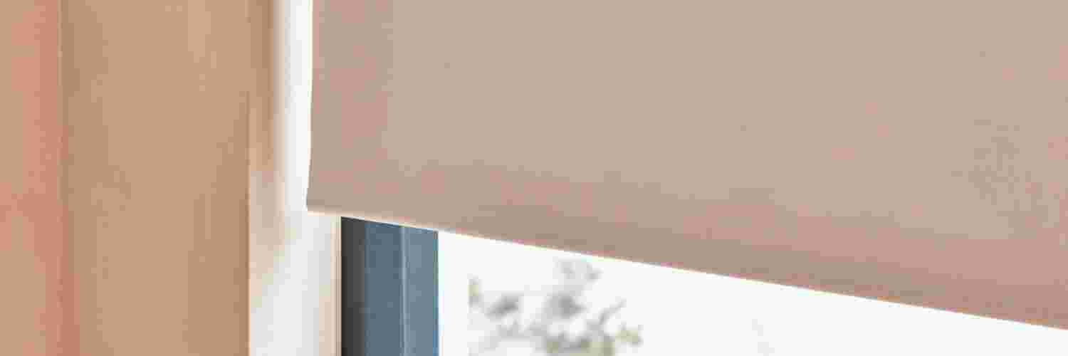 Lichtroze rolgordijn gedeeltelijk neergelaten voor een raam, met zacht licht dat langs de zijkanten binnenvalt.