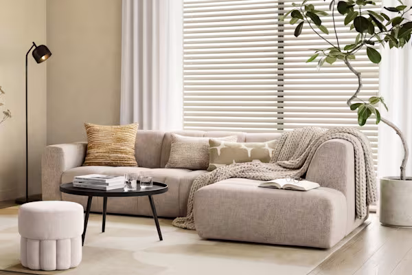 Lichte woonkamer met beige hoekbank, ronde salontafel, staande lamp, jaloezieën en groene kamerplant bij het raam.
