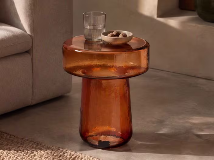 Amberkleurig glazen bijzettafel in paddenstoelvorm met een glas en schaaltje noten erop, naast een beige bank.