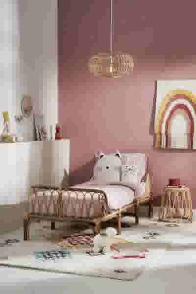 Roze meisjeskamer met een rotan bed, kattenkussen, regenboog wanddecoratie, hanglamp en een wit tapijt met knuffel.
