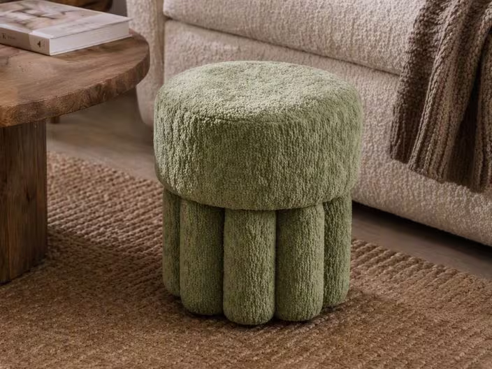 Groene pluche poef met geribbelde basis op een juten vloerkleed, naast een houten salontafel en beige bank.