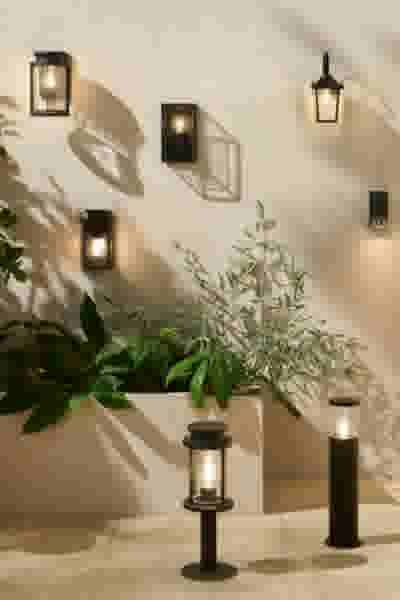 Zwarte buitenlampen op een beige muur en vloer, naast een plantenbak met groene planten en twee staande lantaarns.