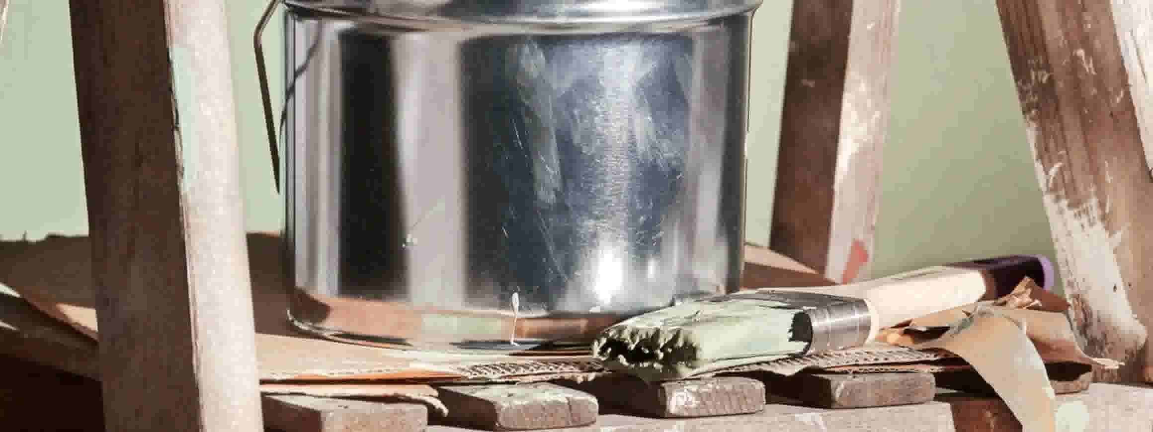 Metalen melkkan op een houten plank met een groene fles en keukengerei tegen een lichtgroene muur.