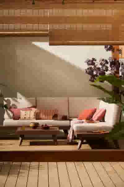 Gezellige tuin met beige hoekbank, kleurrijke kussens en bamboe rolgordijnen, versierd met paarse bloemen in warm zonlicht.