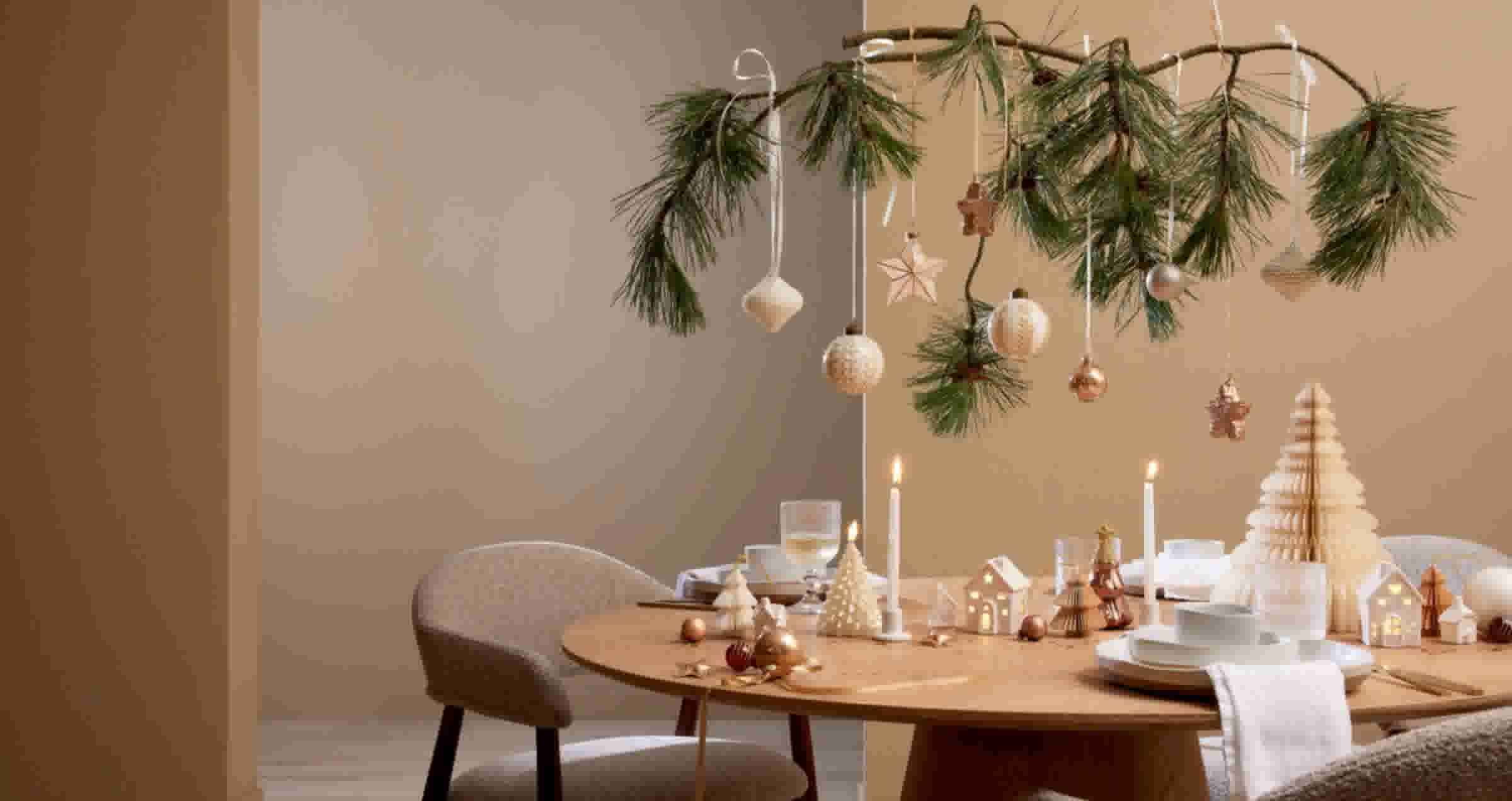 Een feestelijk gedekte ronde tafel in naturel tinten met kaarsen, servies en kleine kerstdecoraties. Boven de tafel hangt een dennentak met subtiele kerstballen in wit en koper.