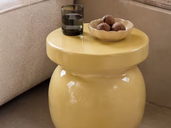 Geel glanzend keramiek bijzettafeltje met een glas water en een schaaltje walnoten, naast een beige bank.