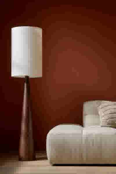 Staande lamp met houten voet en witte kap naast een crèmekleurige bank, tegen een terracotta rode muur.