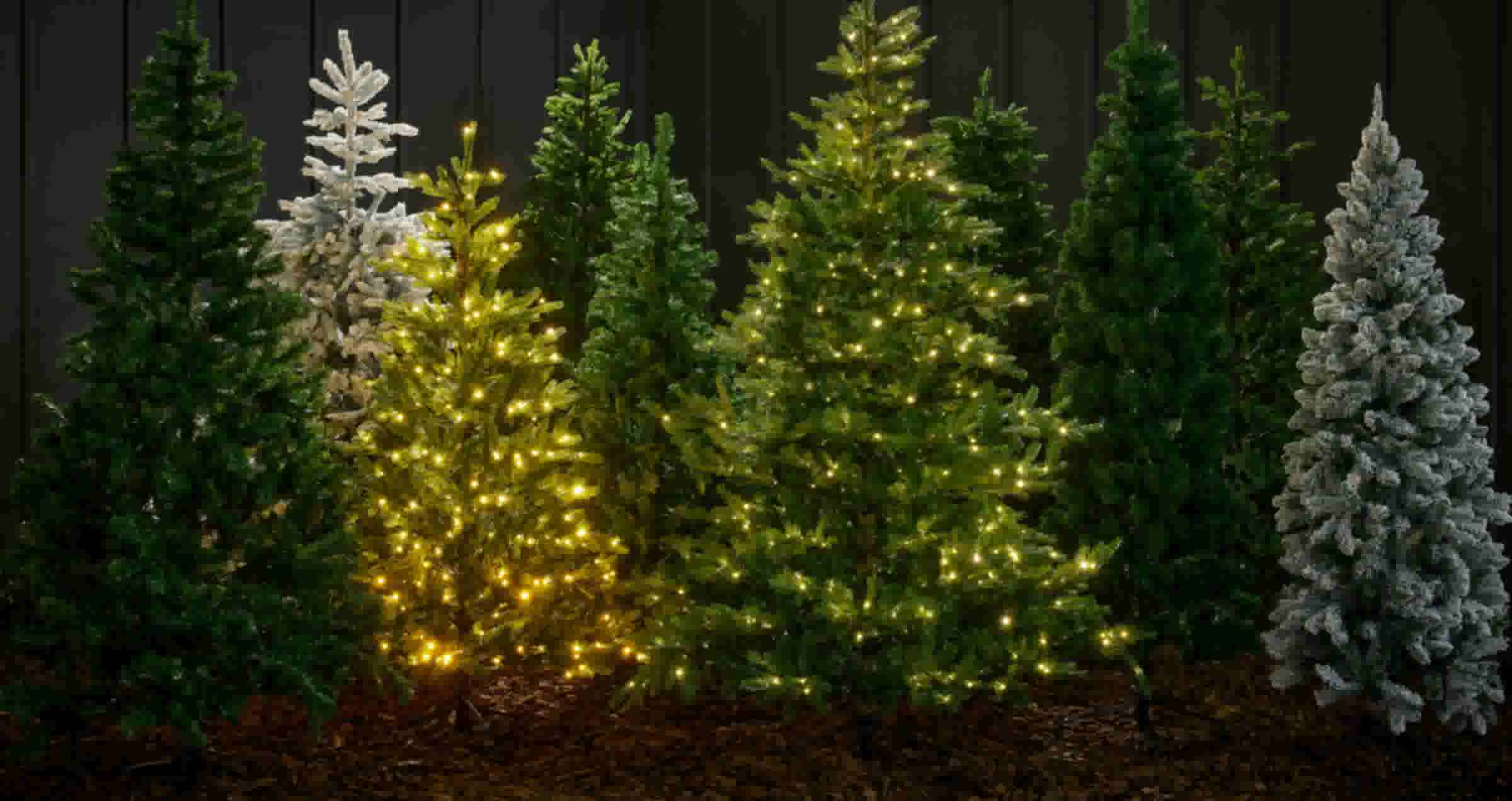 Rij kunstkerstbomen in verschillende groentinten, sommige met witte sneeuweffecten en andere versierd met warme lichtjes.