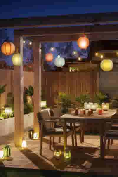 Sfeervolle tuinpergola bij avond met kleurrijke lampionnen, lichtjes en lantaarns rond een houten eettafel met stoelen.